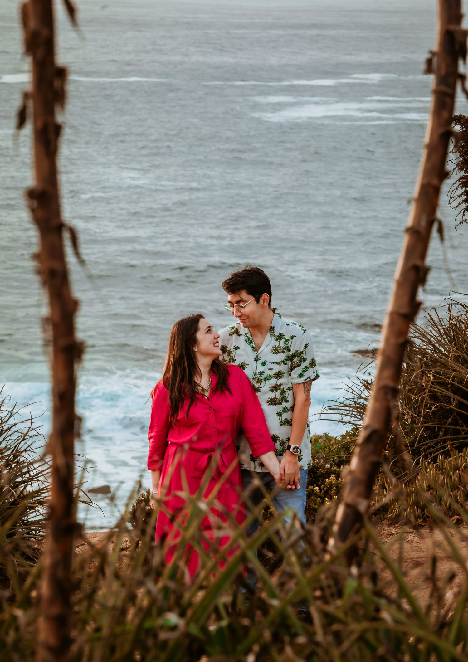 Couple gazing at each other on a coastal cliff with ocean waves in the background, framed by tropical plants.