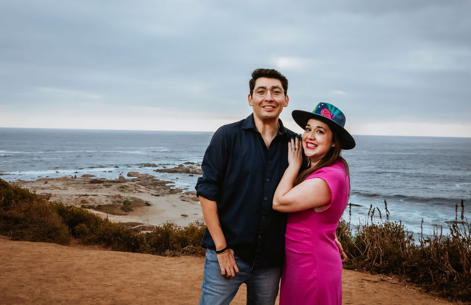 A couple smiles together on a coastal cliff overlooking the ocean, woman in pink dress and man in navy shirt.