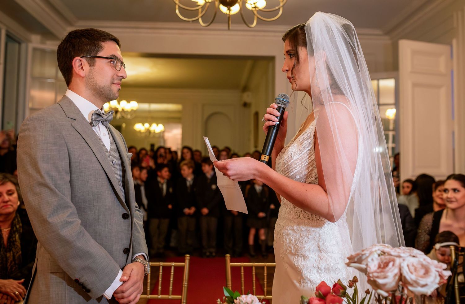 Bride reads wedding vows to groom at elegant indoor ceremony, wearing white gown and veil, holding microphone and bouquet.