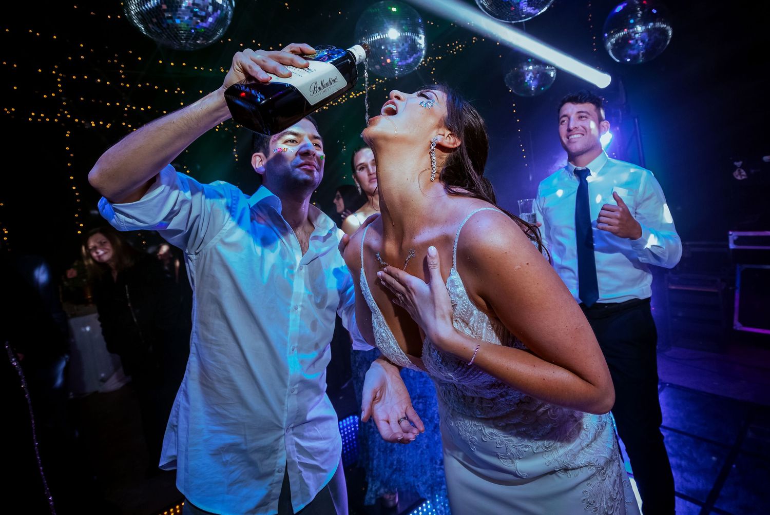 A groom pours champagne into a brides mouth on the dance floor under disco ball lights at a wedding reception.