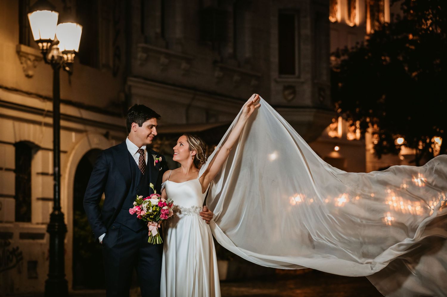 Bride and groom gazing at each other as wedding veil billows dramatically under city lights.