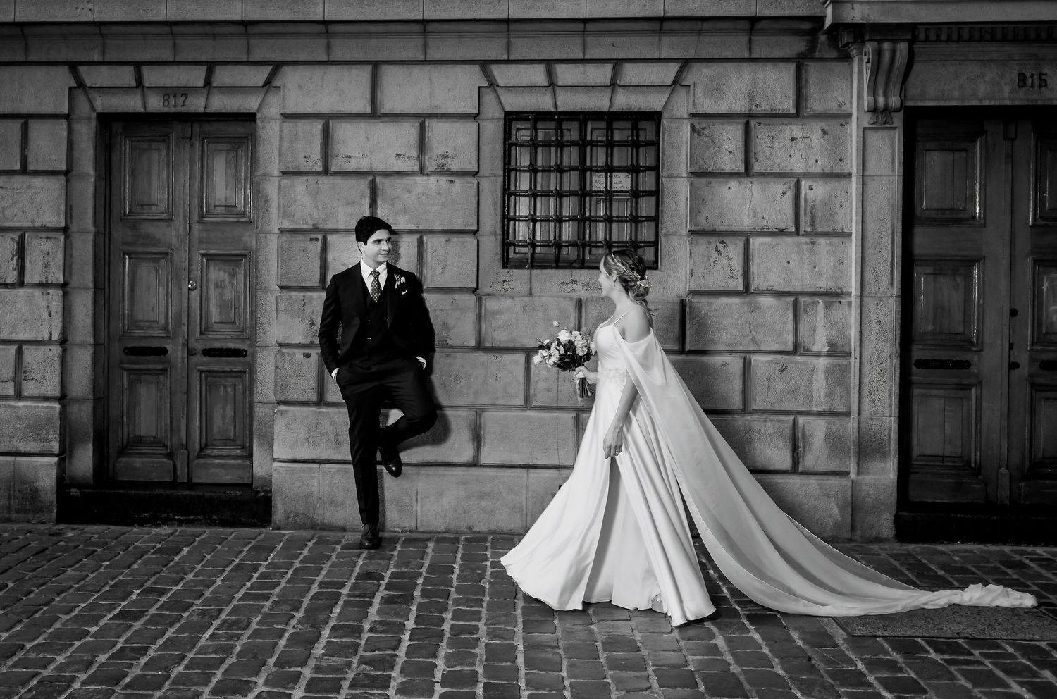 Black and white photo of groom leaning on wall as bride approaches on cobblestone street.