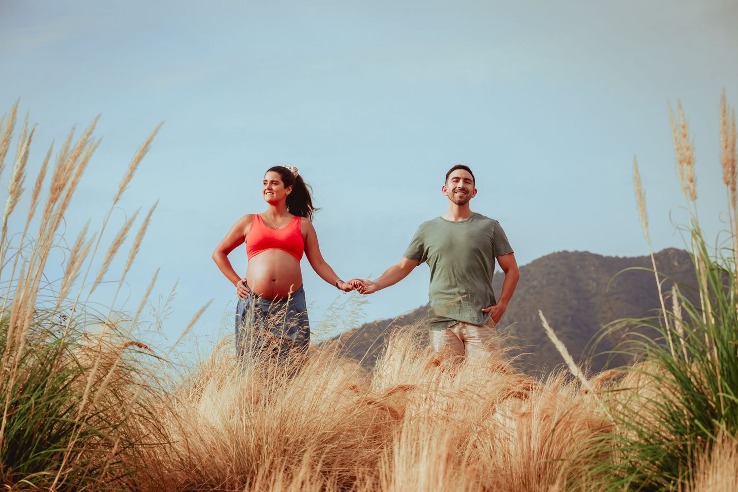 Happy pregnant couple holding hands walking through tall golden grass with mountain backdrop and blue sky.