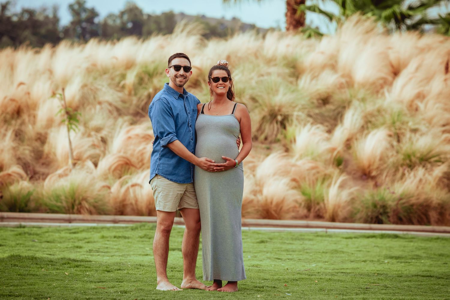 Couple posing on green lawn, man holding pregnant woman's belly with pampas grass background.