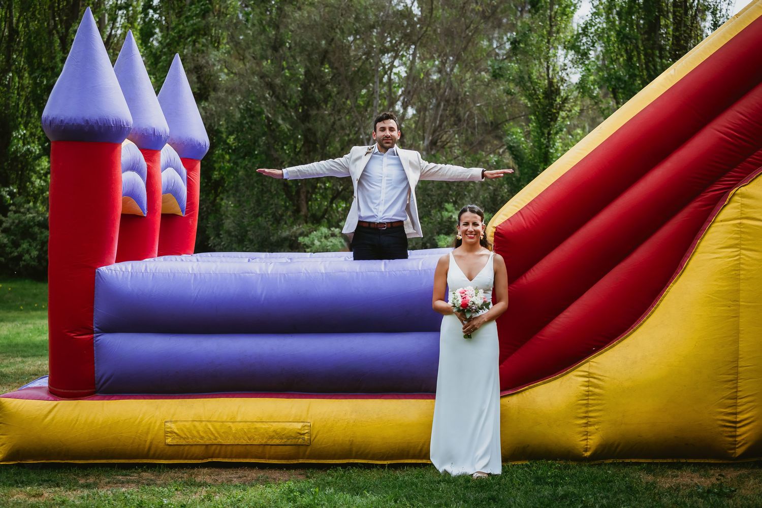 Bride and groom posing joyfully in front of a colorful inflatable bounce castle at their outdoor wedding reception.