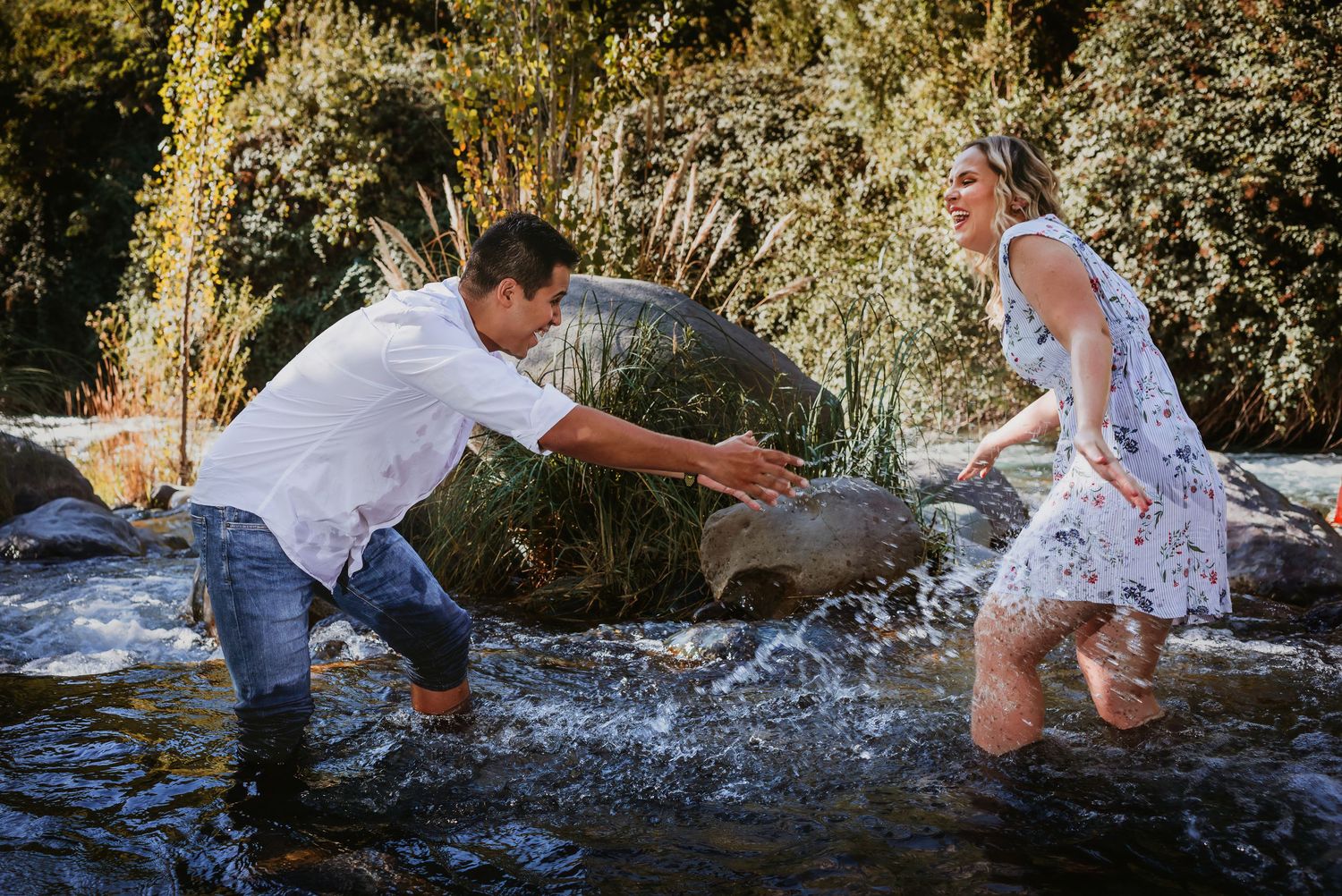 Couple playing in a rocky stream, man reaching out to woman in floral dress, surrounded by lush greenery.