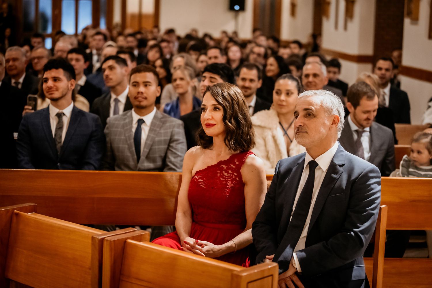 Guests seated in church pews during a wedding ceremony, woman in red dress beside man in suit.