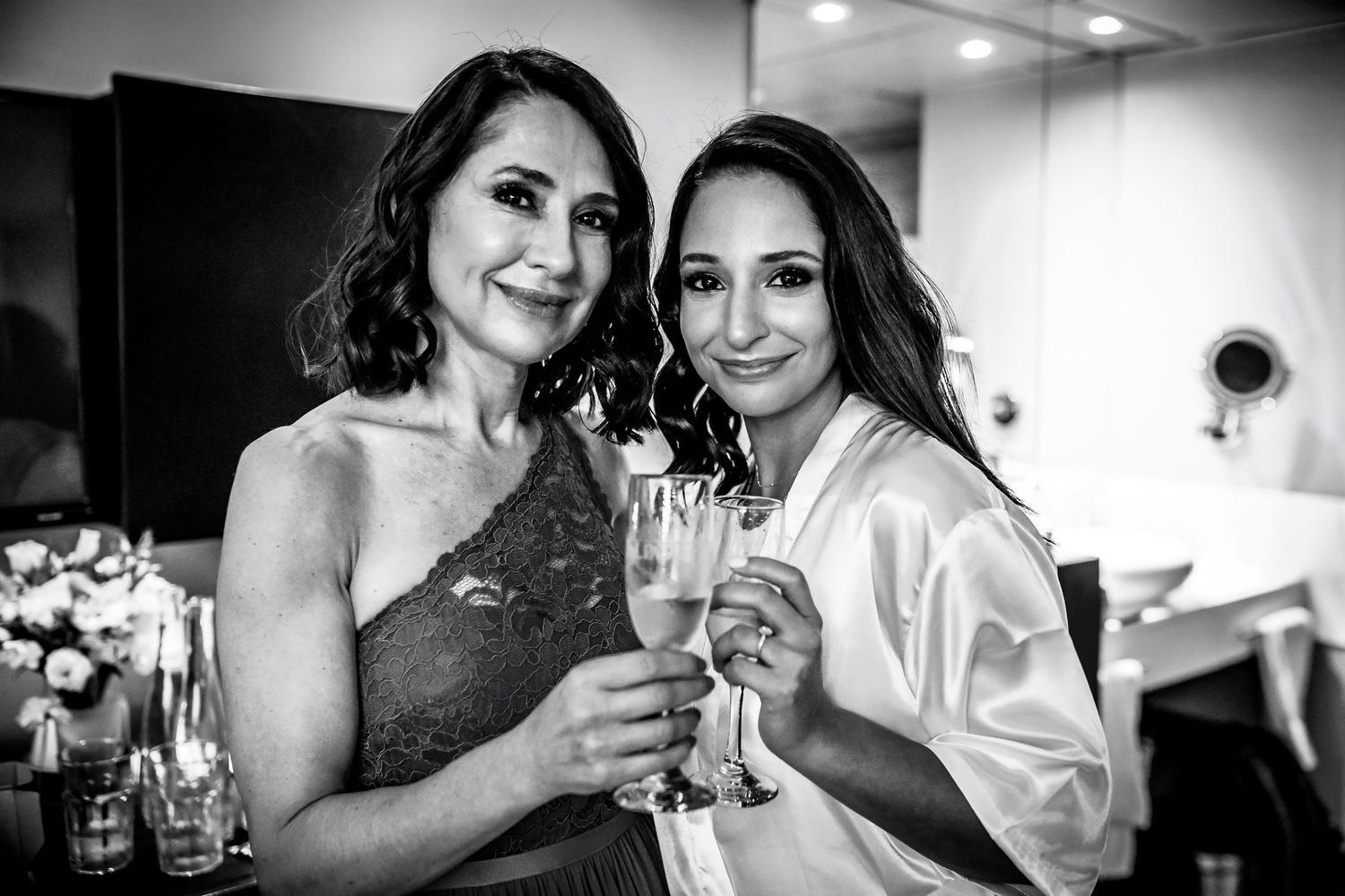 Two women toast with champagne glasses in a black and white photo, one in a robe, celebrating a wedding day.