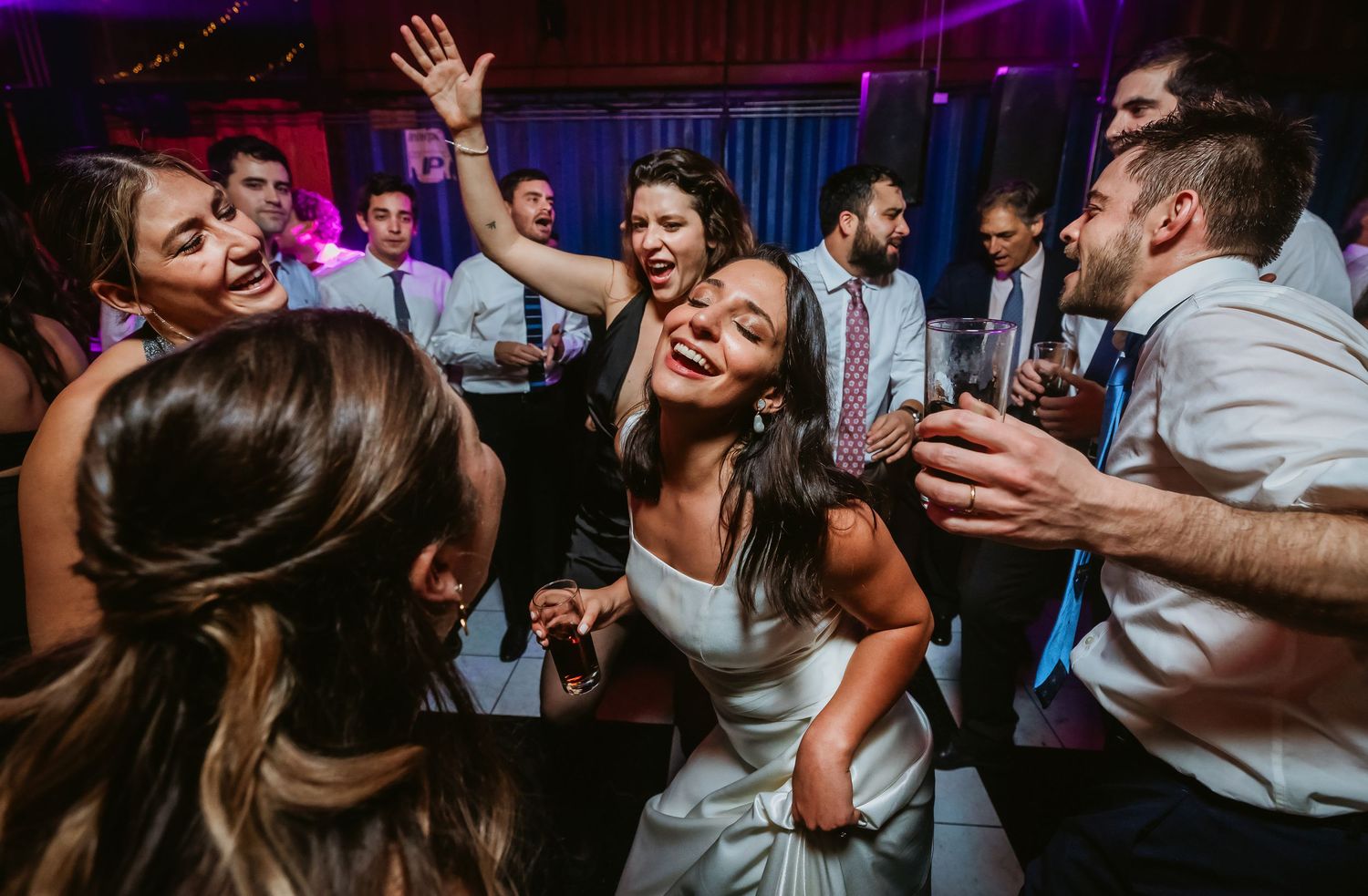 Bride dancing joyfully with wedding guests at lively reception party with colorful lights.