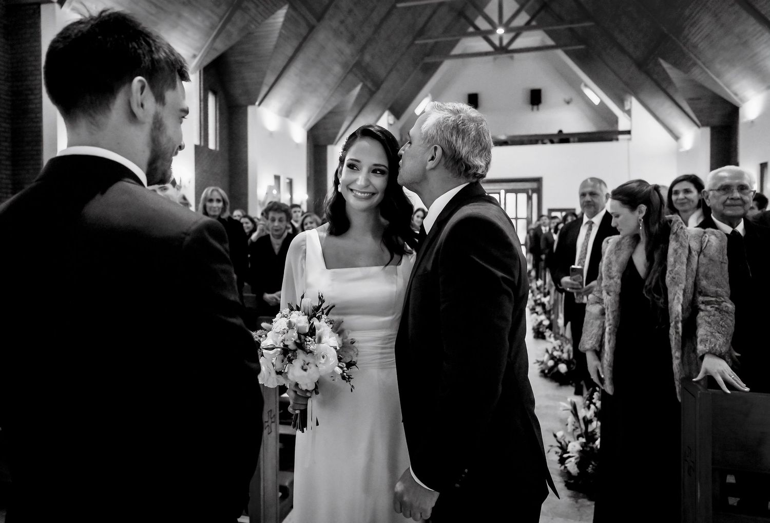 Black and white wedding photo of bride receiving a kiss from father at church altar while holding bouquet.