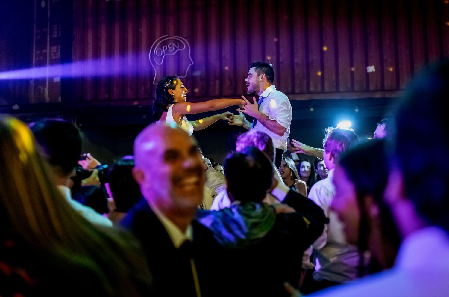 Groom lifted by guests during lively wedding reception celebration with dramatic lighting.