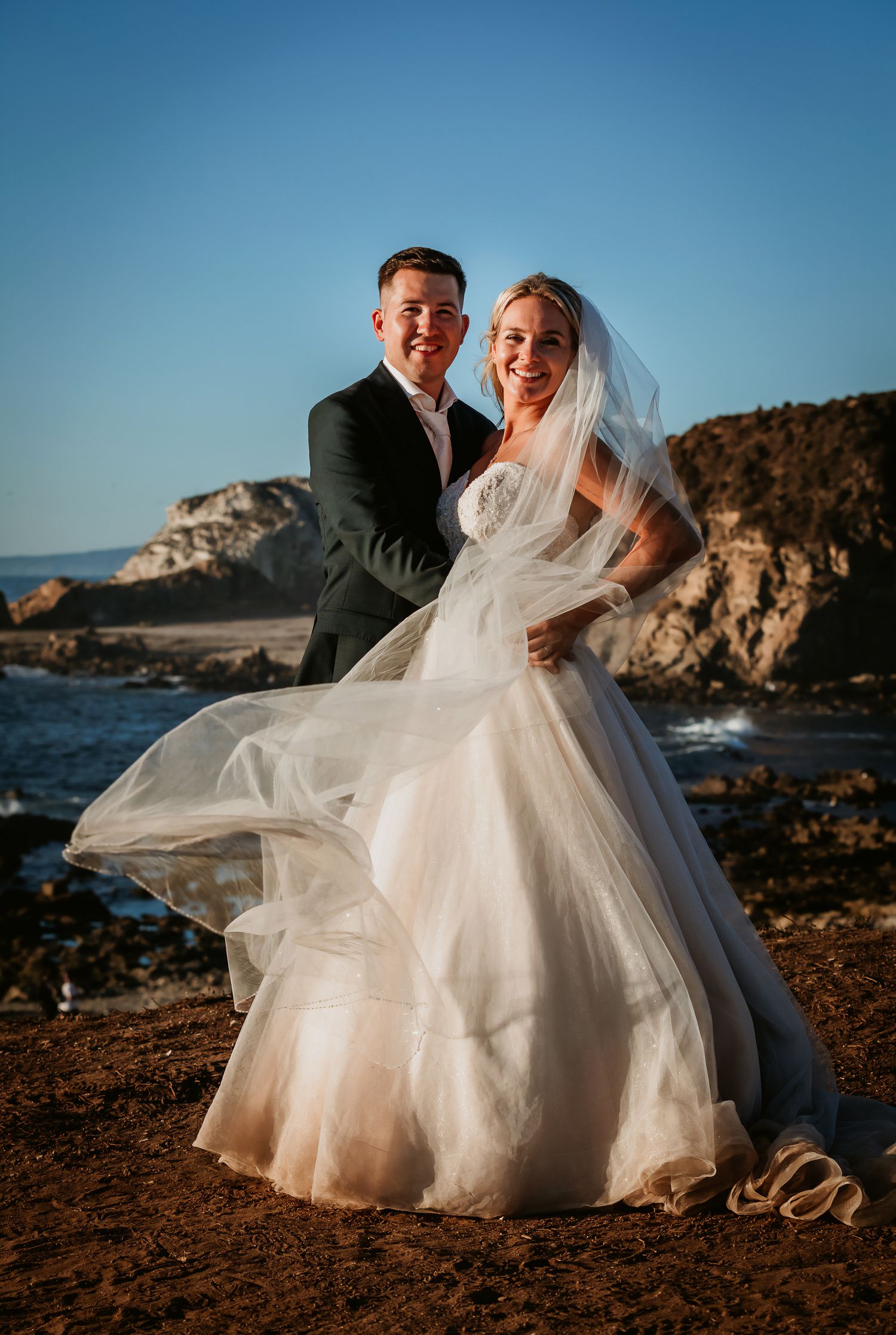 Laughing bride and groom pose together on rocky beach, her white gown and veil flowing in breeze.
