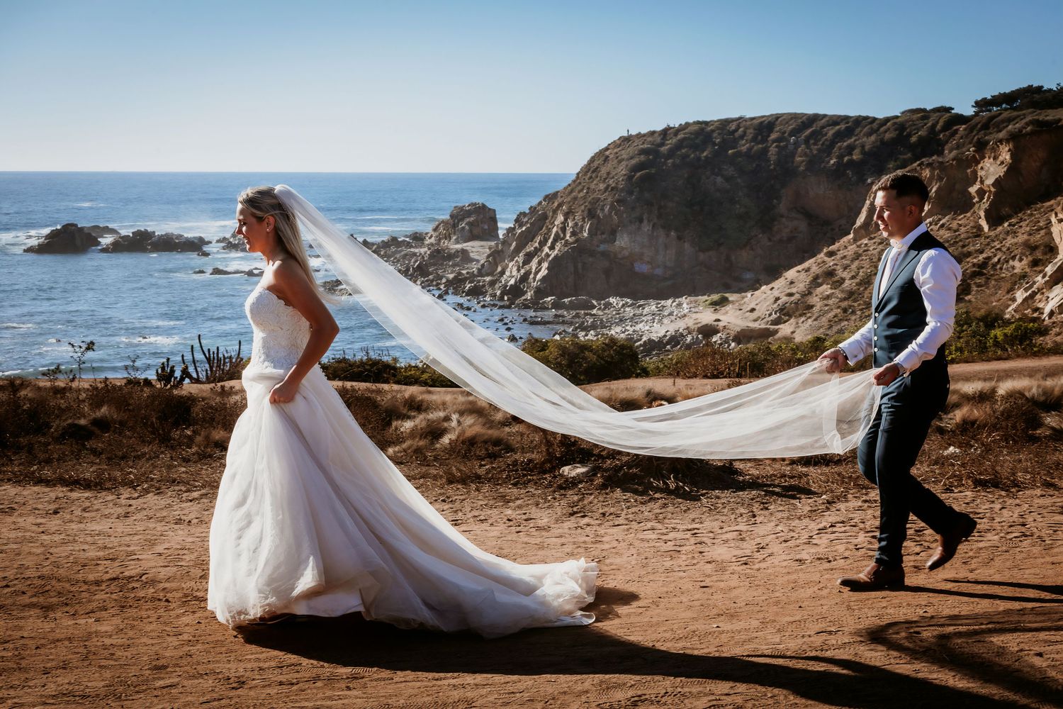 Groom chases the bride's flowing veil along a rugged coastal path with dramatic rocky cliffs and ocean views.