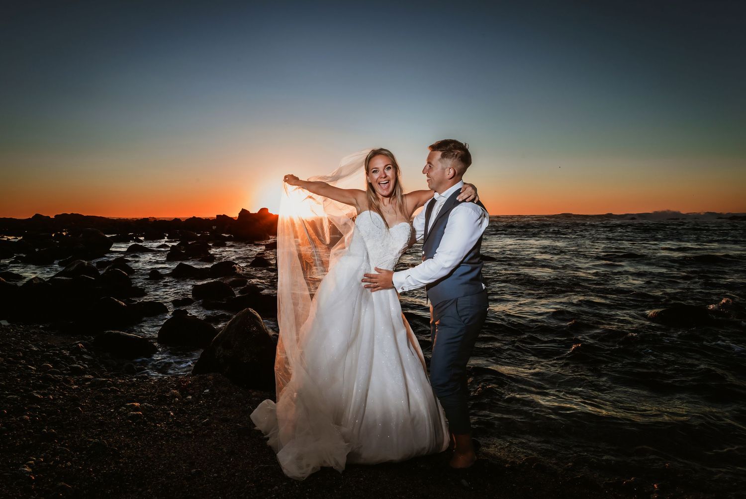 Bride and groom dancing playfully on rocky shoreline with glowing orange sunset behind.