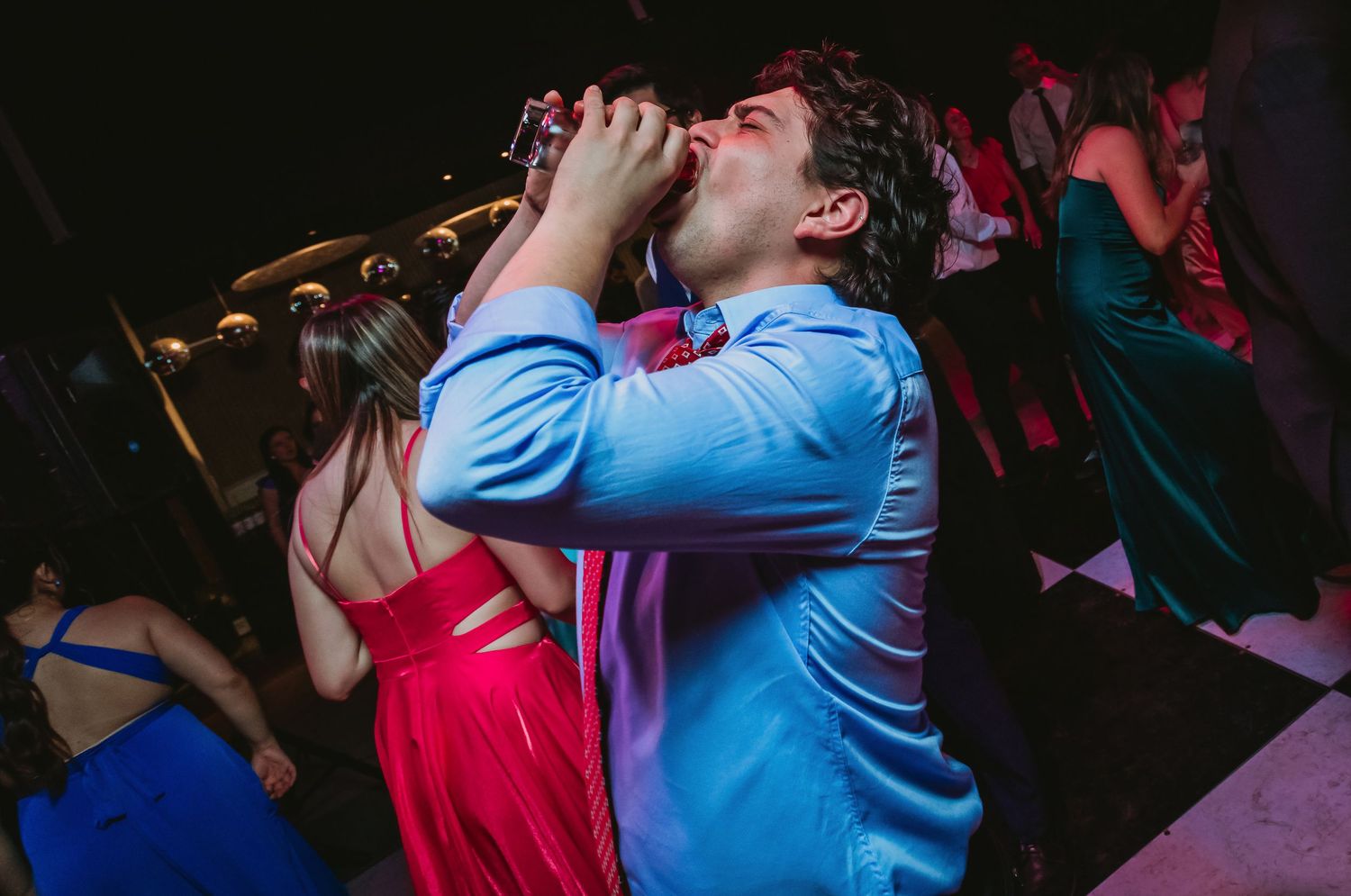 Man in blue suit drinks at lively wedding reception dance floor with guests in background.