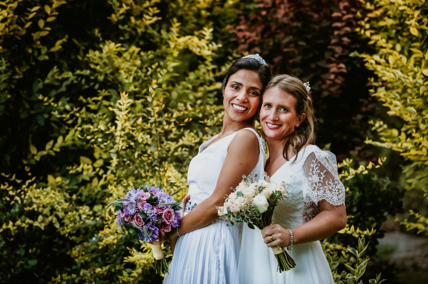 Two brides smile joyfully holding bouquets in front of lush green foliage on their wedding day.