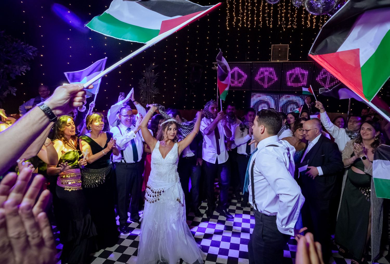 Bride and groom dancing as guests wave Palestinian flags at a lively wedding reception.