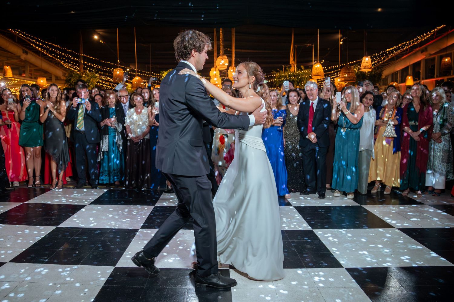 Bride and groom sharing romantic first dance on black and white checkered floor surrounded by guests.