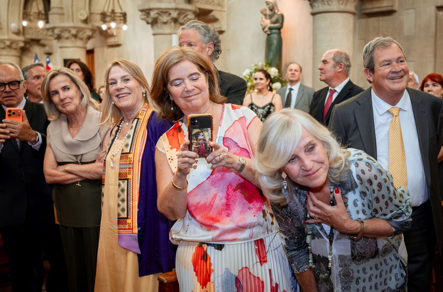 Elegantly dressed guests taking selfies and socializing inside ornate church wedding ceremony.