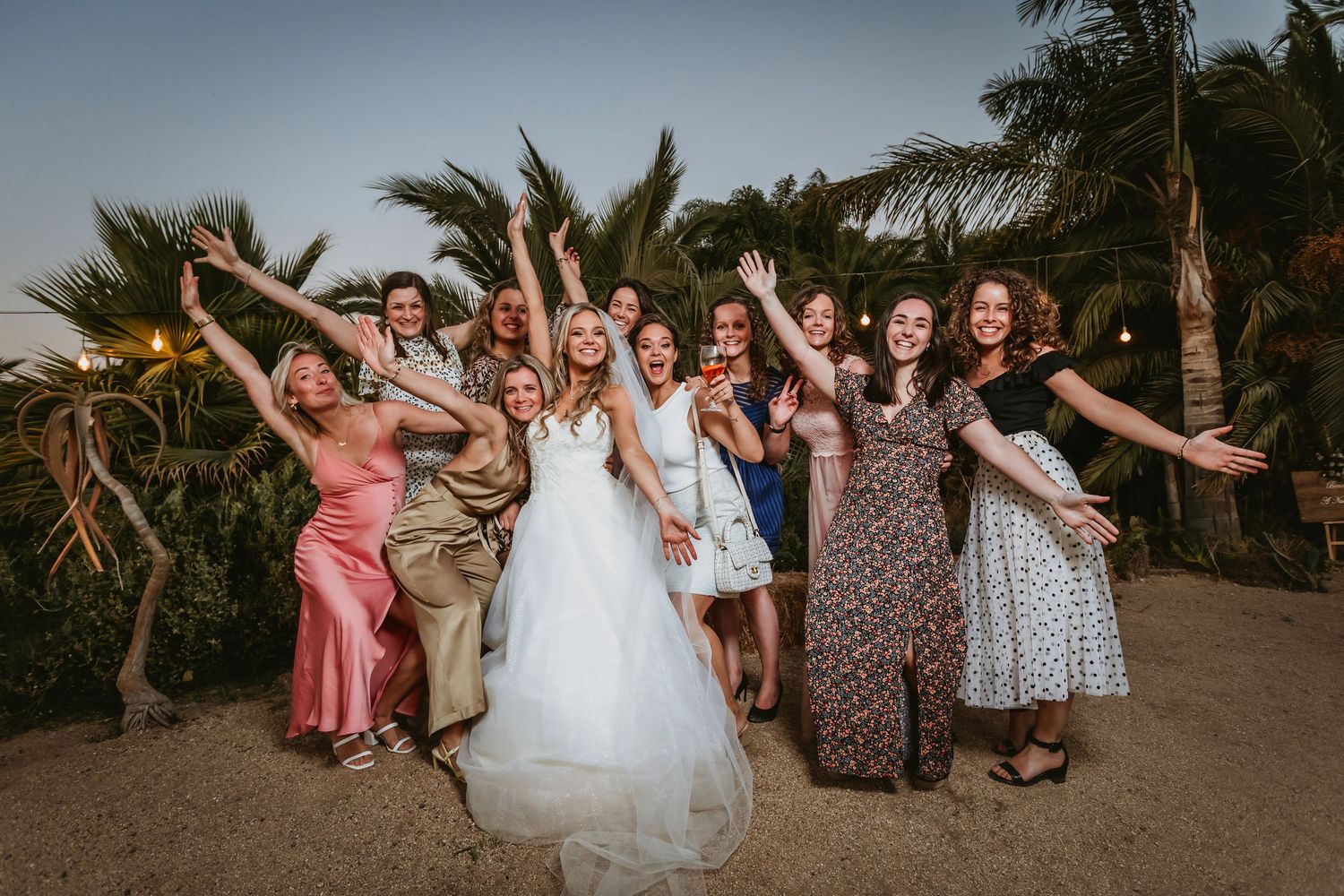 Bride celebrates with group of bridesmaids raising arms joyfully outdoors at tropical wedding reception.
