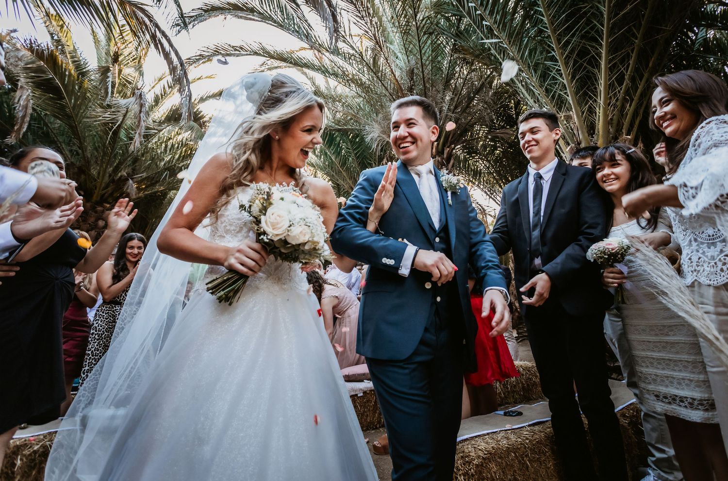 Joyful newlyweds walk through confetti shower among palm trees, bride in white gown holding bouquet.