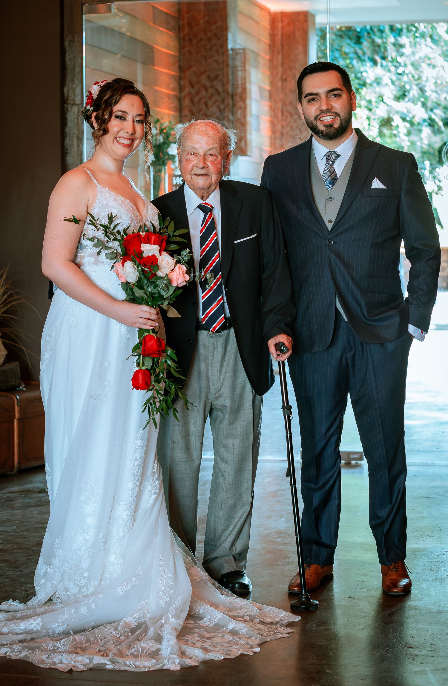 Bride and groom posing with elderly gentleman using a cane at an outdoor wedding venue.