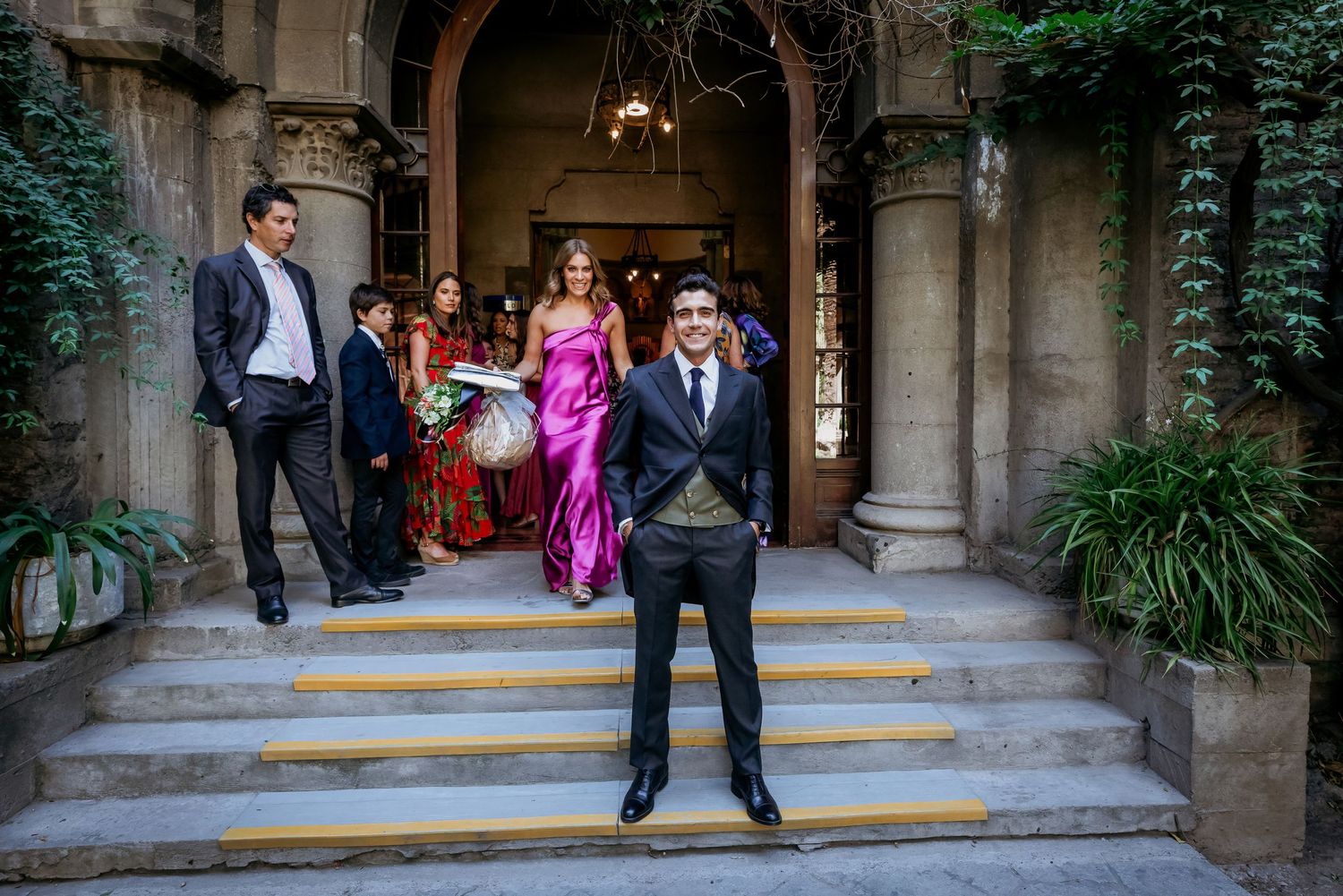 Groom posing on ornate building steps with wedding party emerging from decorated entrance.
