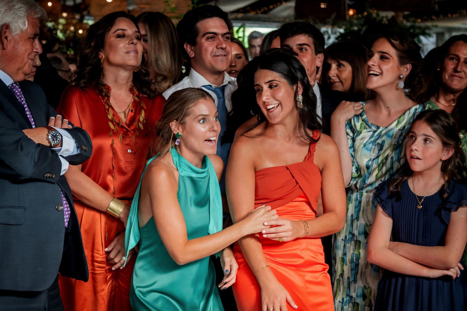 Guests laughing and celebrating at a wedding reception, women wearing colorful orange and teal formal dresses.