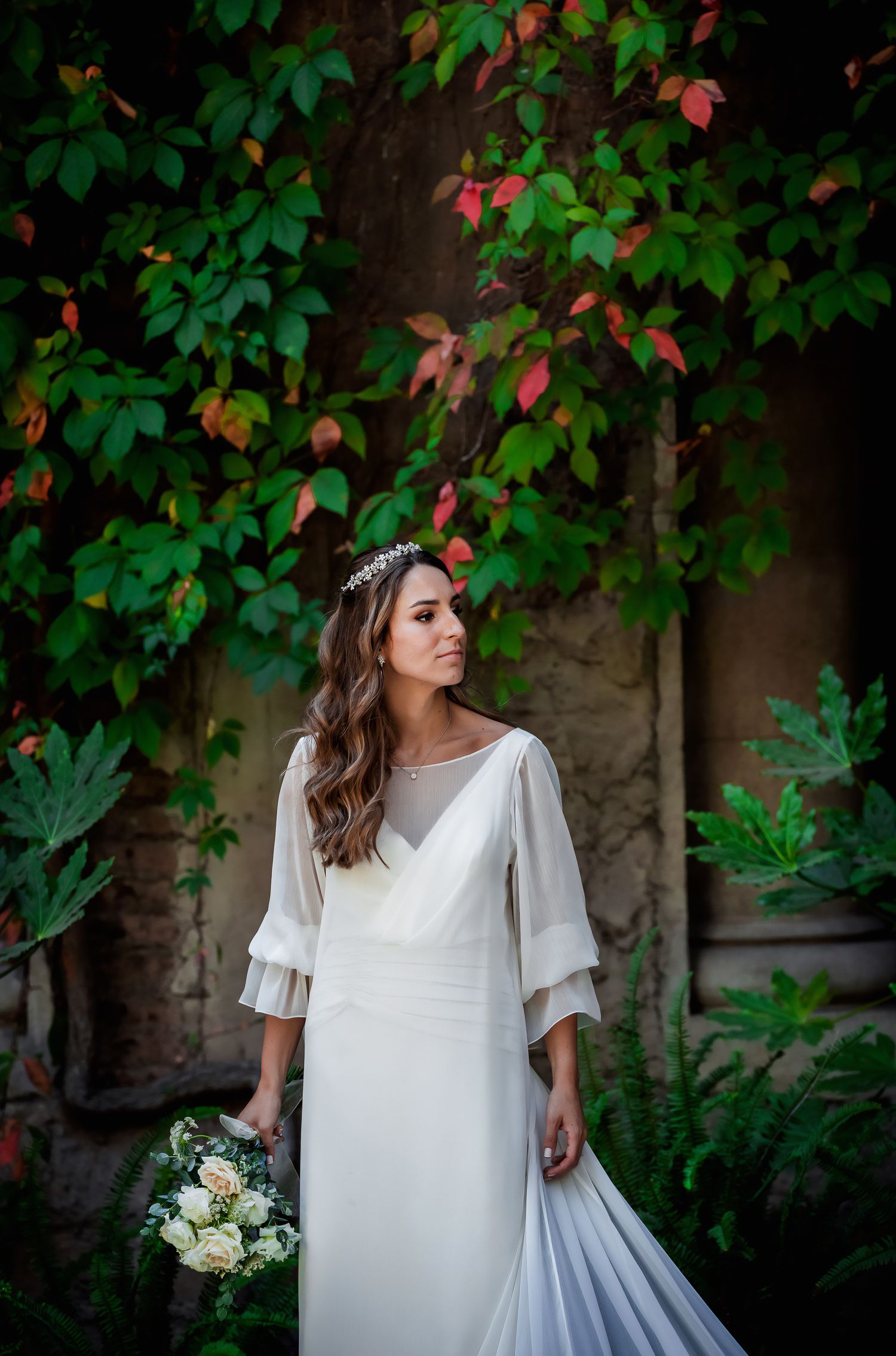 Elegant bride in white gown with tiara holding bouquet standing before colorful autumn ivy wall.