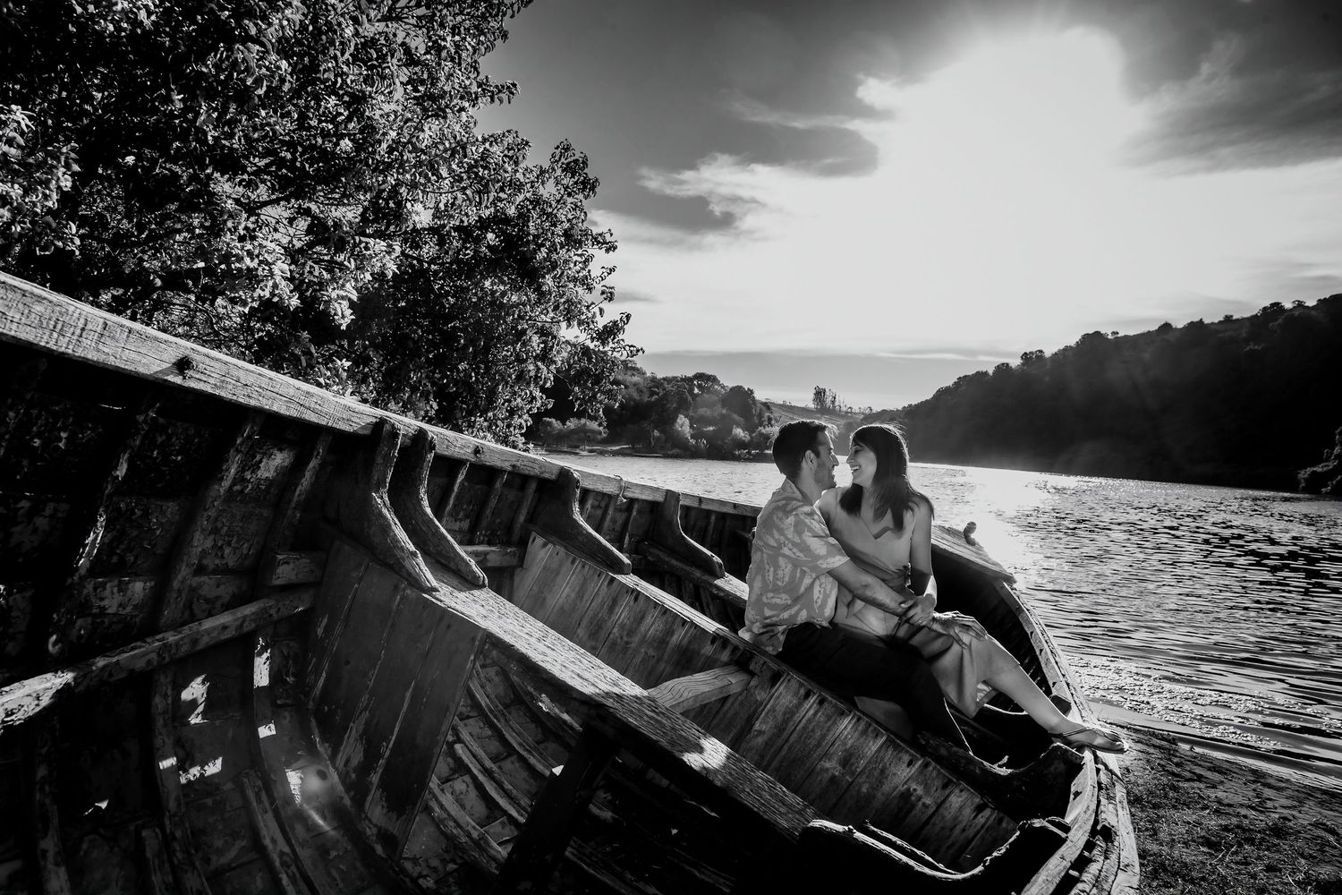 Black and white photo of a romantic couple sitting in a weathered boat by a river.