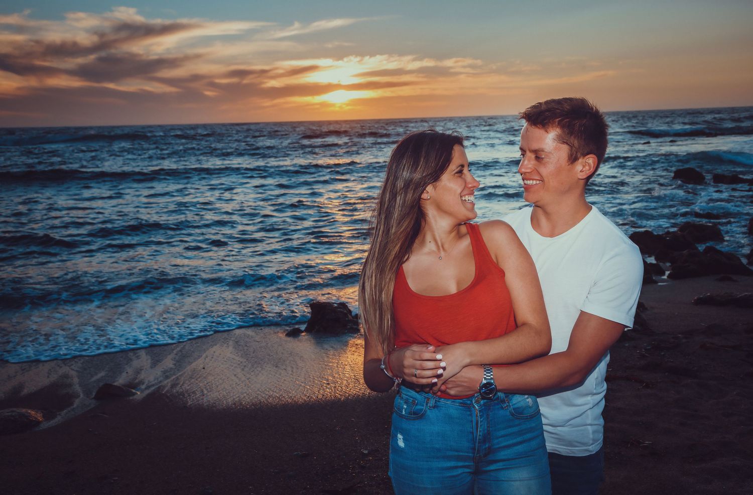 Loving couple embracing on a sandy beach with colorful sunset sky and ocean waves behind them.