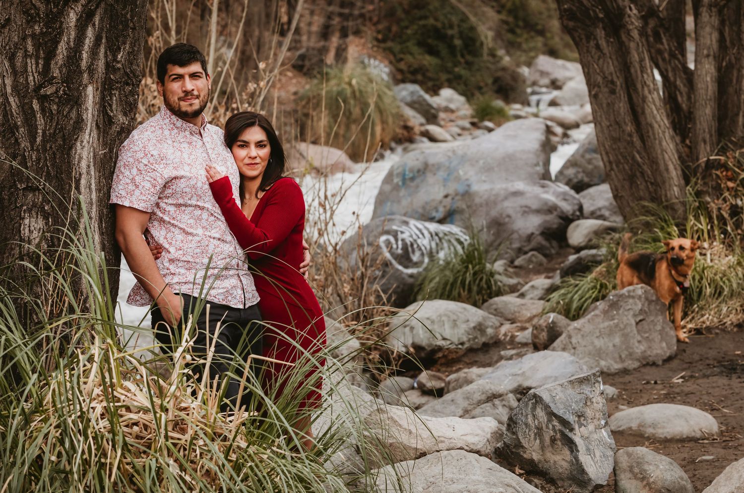 Couple posing seriously among trees and rocks near a stream with their dog nearby.