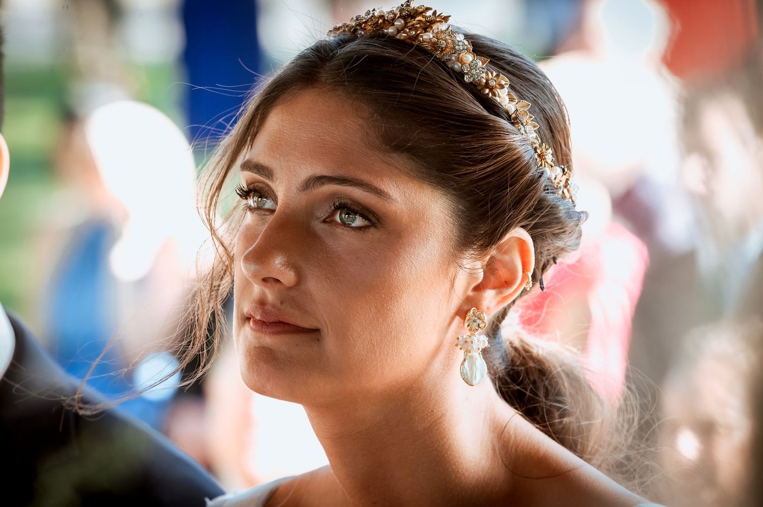Close-up of a beautiful bride wearing a gold floral headpiece and drop earrings at an outdoor wedding ceremony.