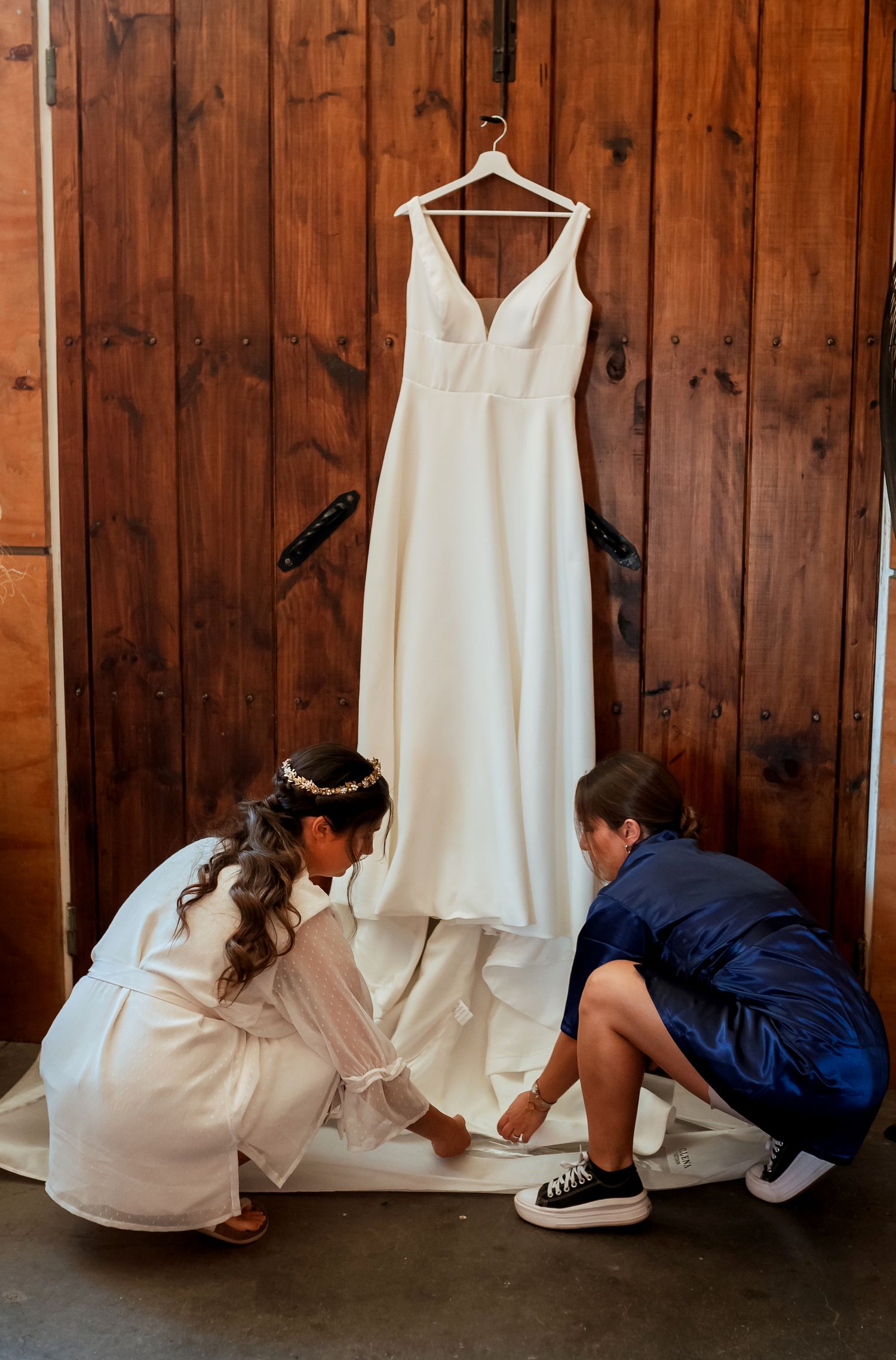 Bride and bridesmaid kneeling to arrange a white wedding dress hanging against a wooden barn wall.