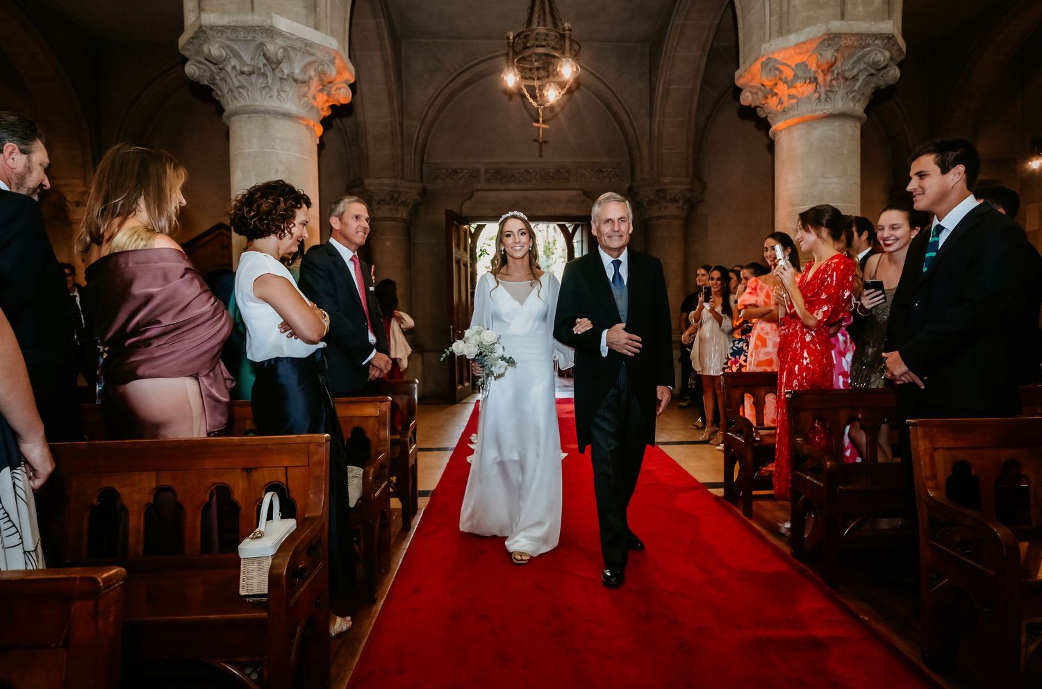 Bride walking down red carpet aisle with escort in ornate church filled with wedding guests.