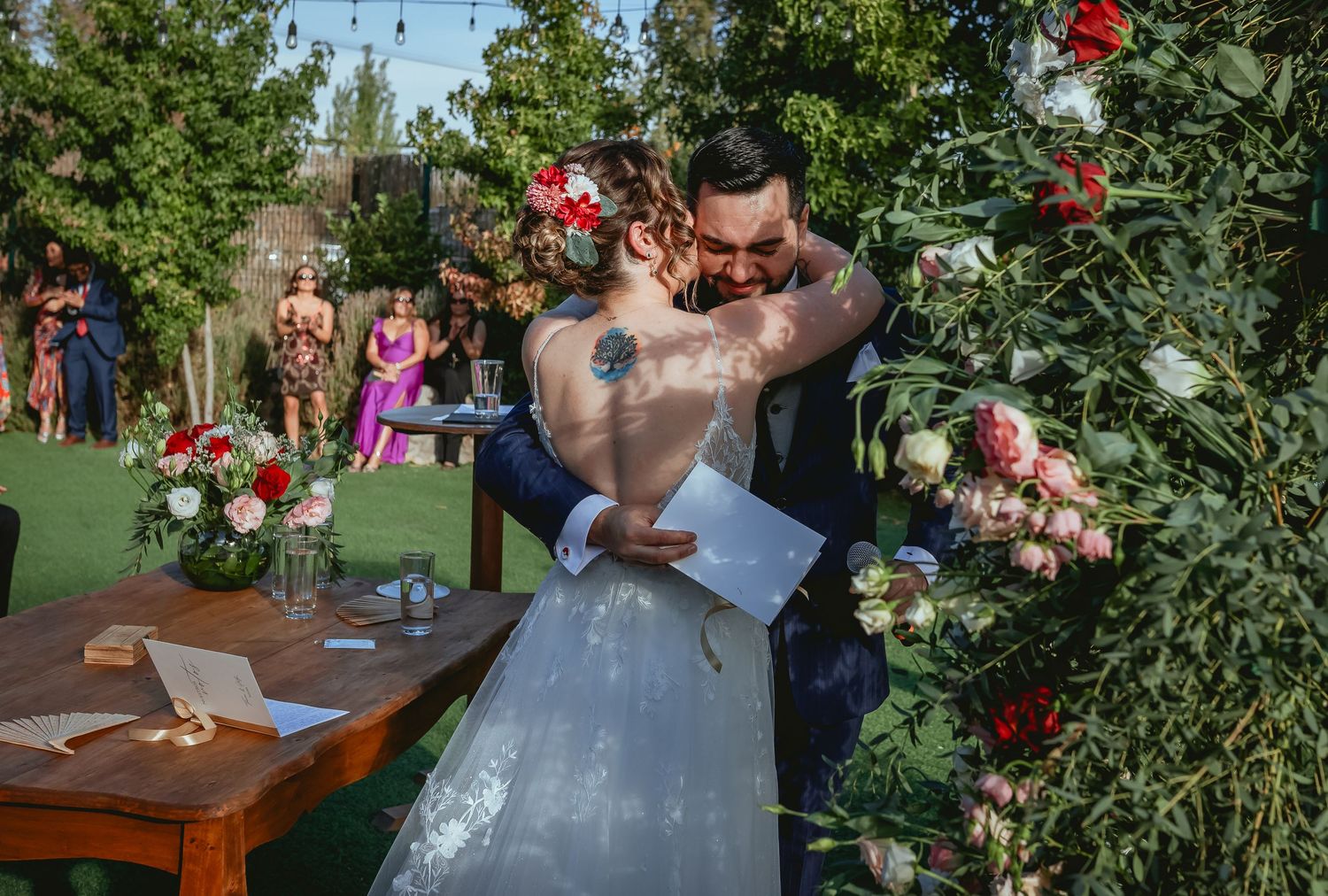 Bride and groom share emotional embrace at floral arch during outdoor garden wedding ceremony.