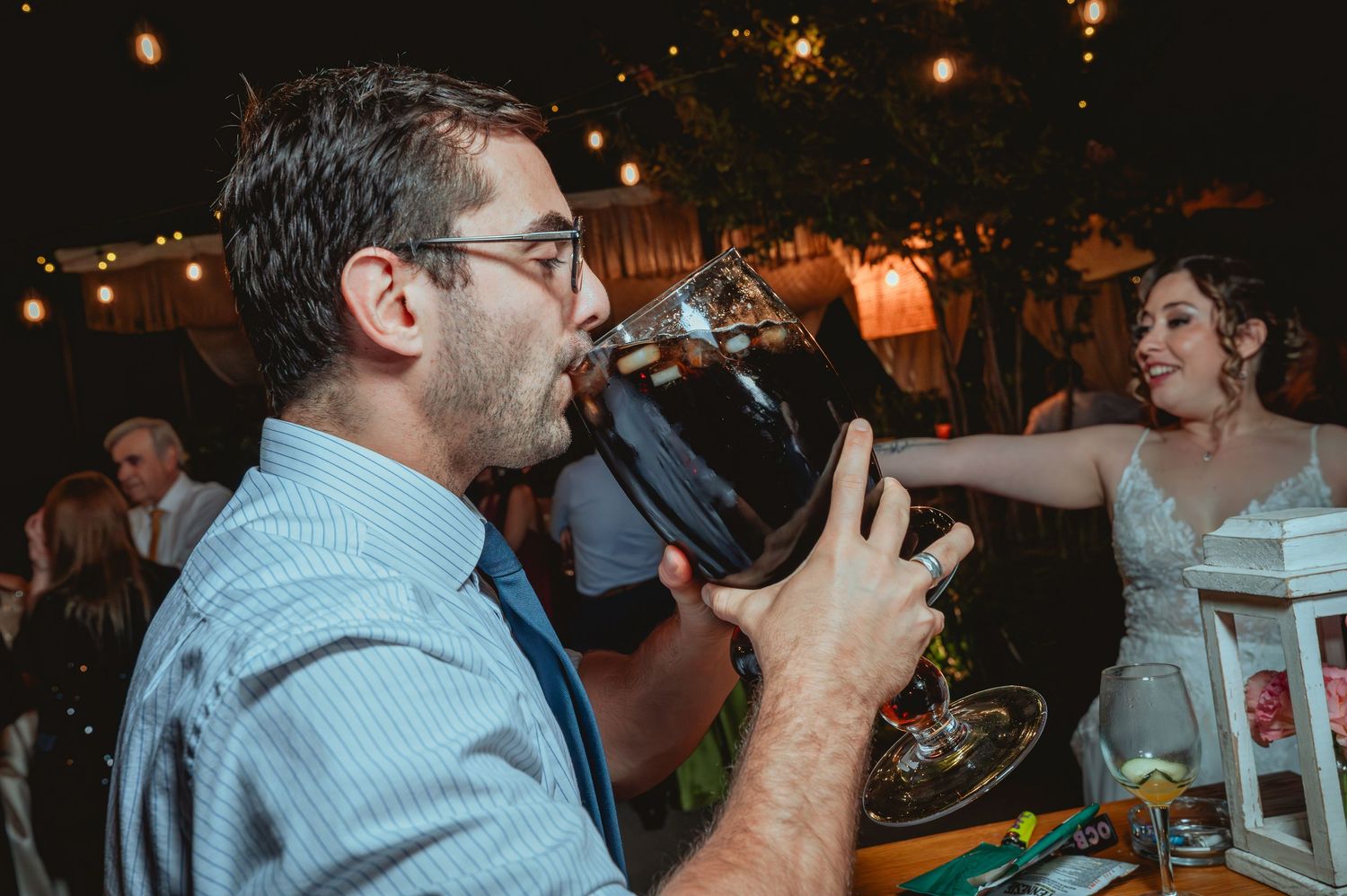 Man drinks from large glass dispenser as bride looks on at wedding reception.