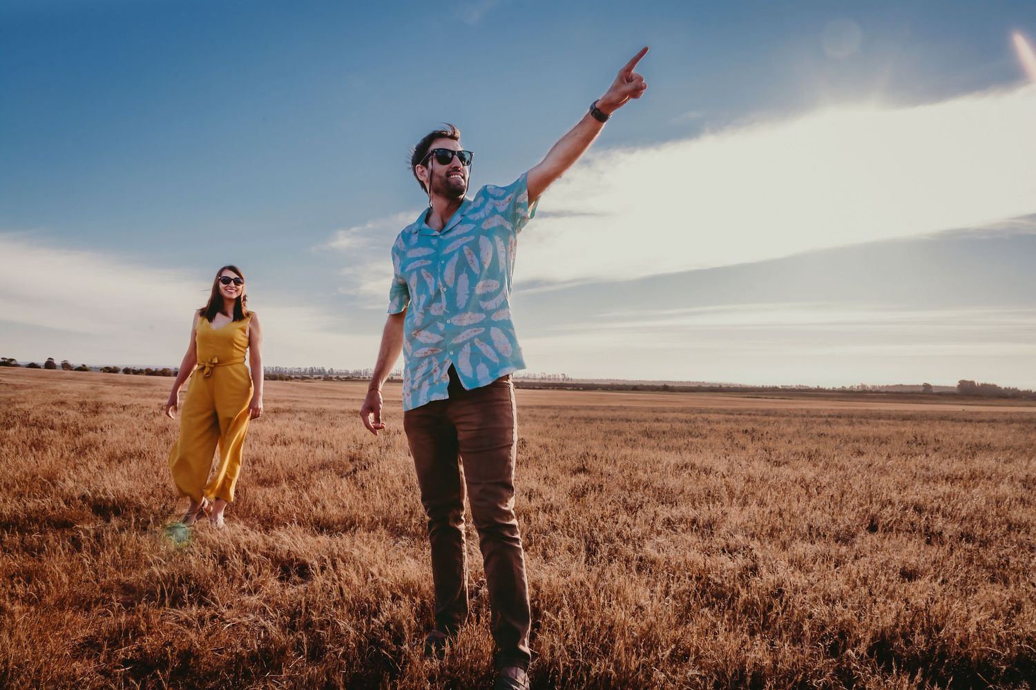 Couple in a golden wheat field, man in blue Hawaiian shirt pointing at sky, woman in yellow dress behind him.