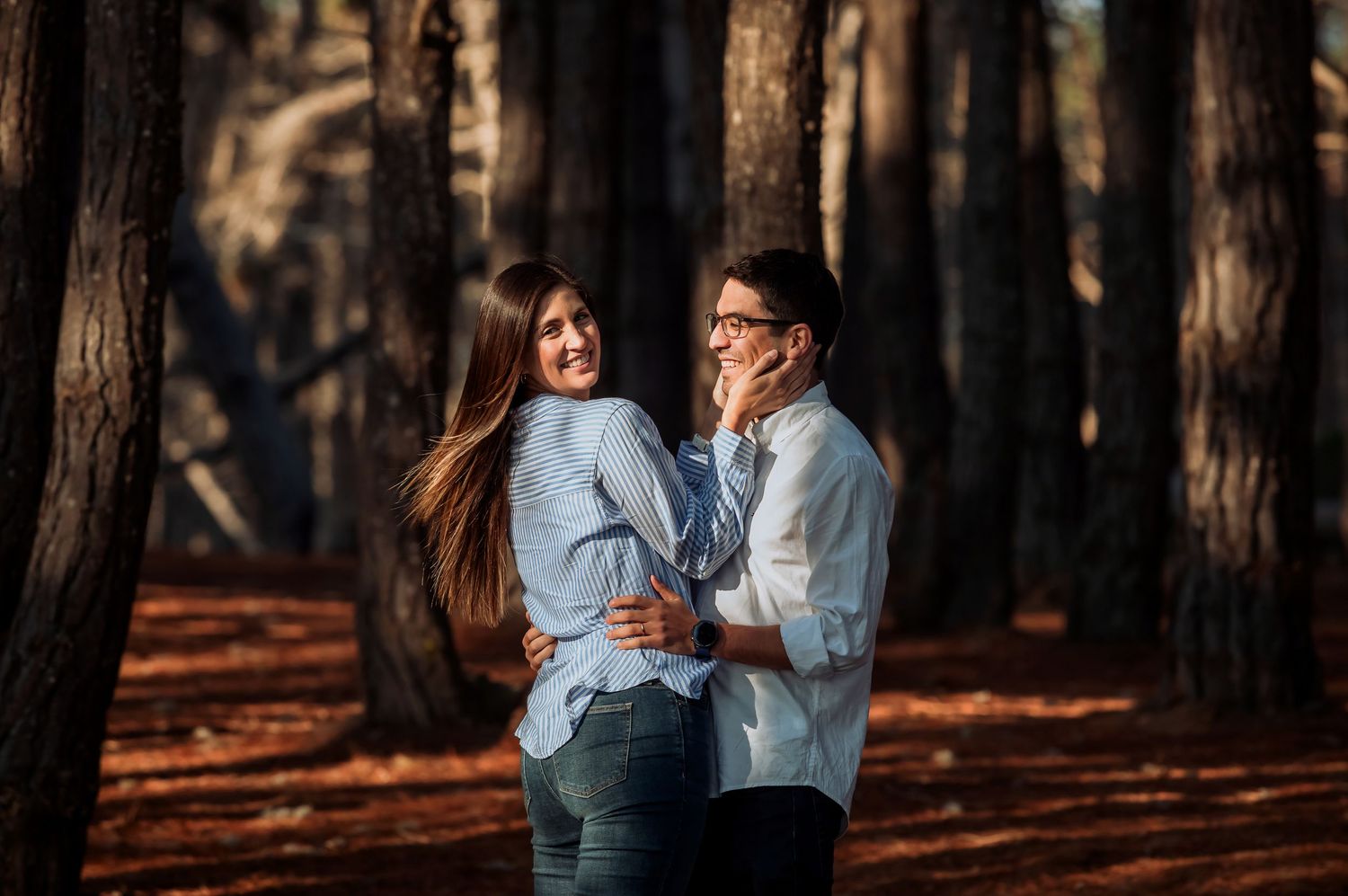A smiling couple embracing and laughing together among tall pine trees in warm sunlight.