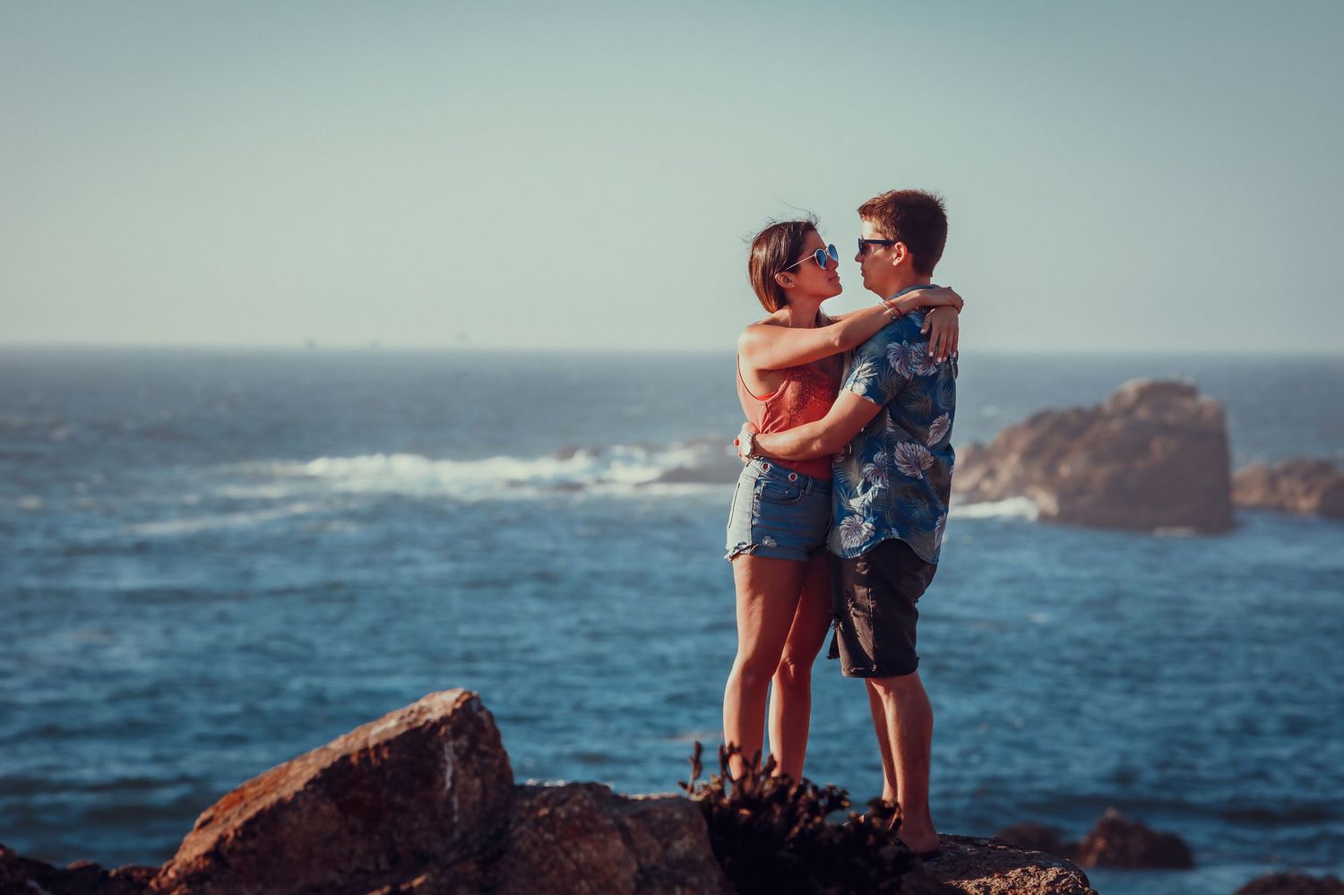 Couple sharing tender moment on rocky coast with blue ocean backdrop.