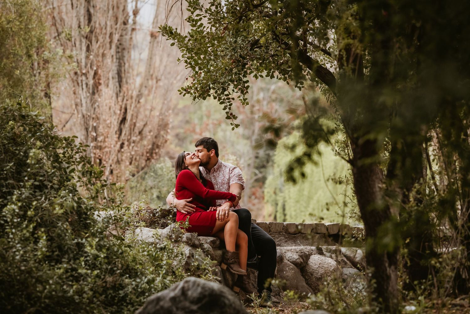 Couple sharing a romantic kiss while sitting on stone wall surrounded by lush green trees in a garden.