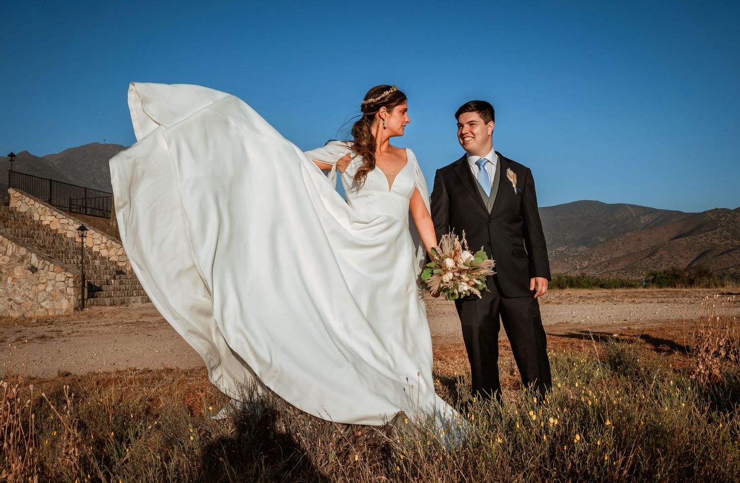 Wedding couple stands together as bride's white gown flows dramatically in the wind with mountains behind.