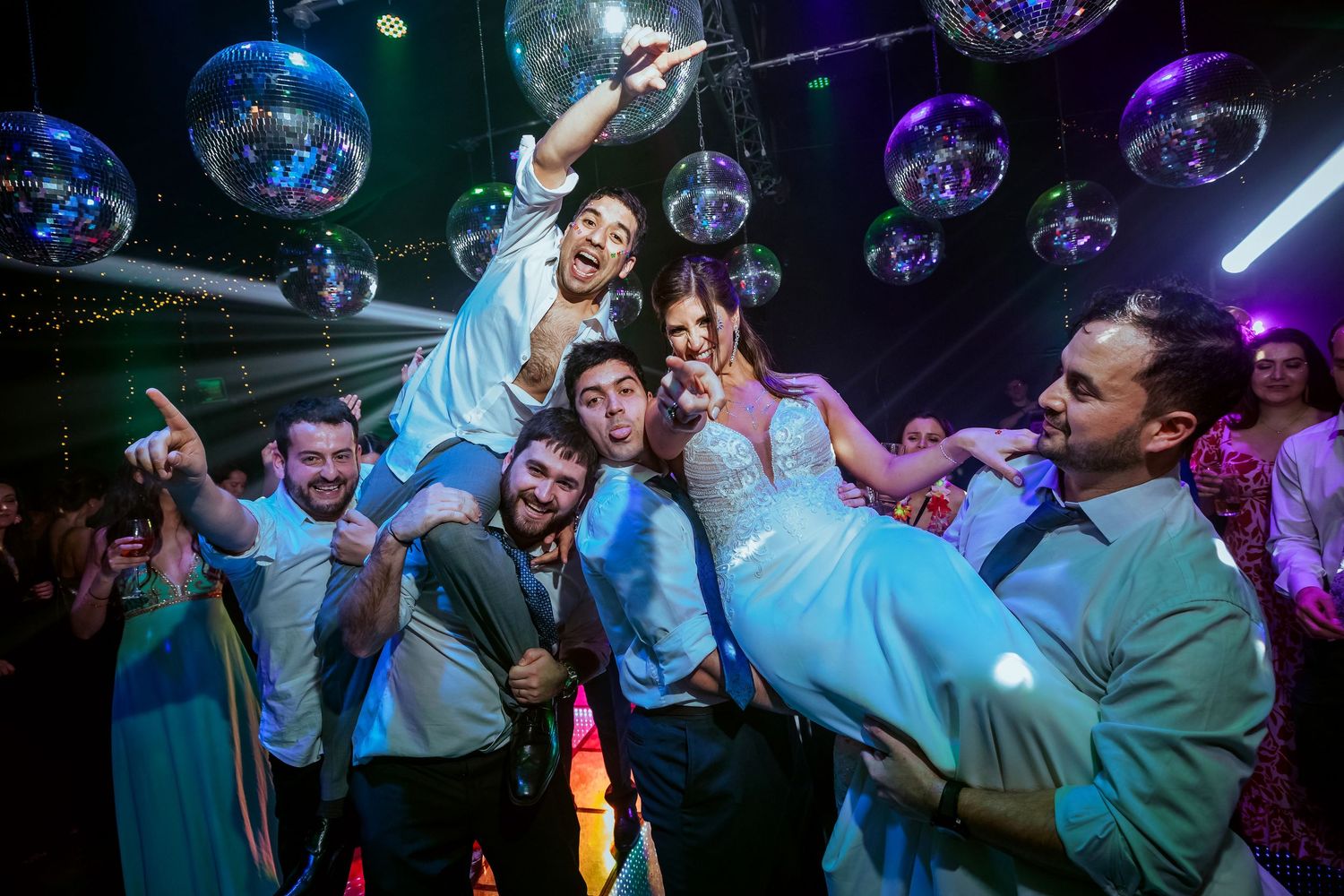 Wedding couple lifted by groomsmen on dance floor with disco balls and colorful lights at reception party.