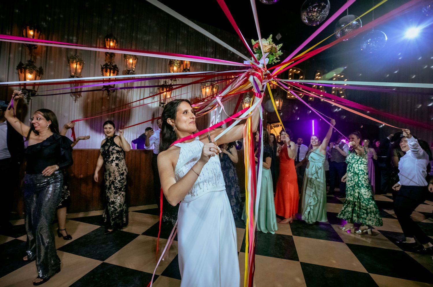 Bride holds colorful ribbon bouquet on checkered dance floor at wedding reception.