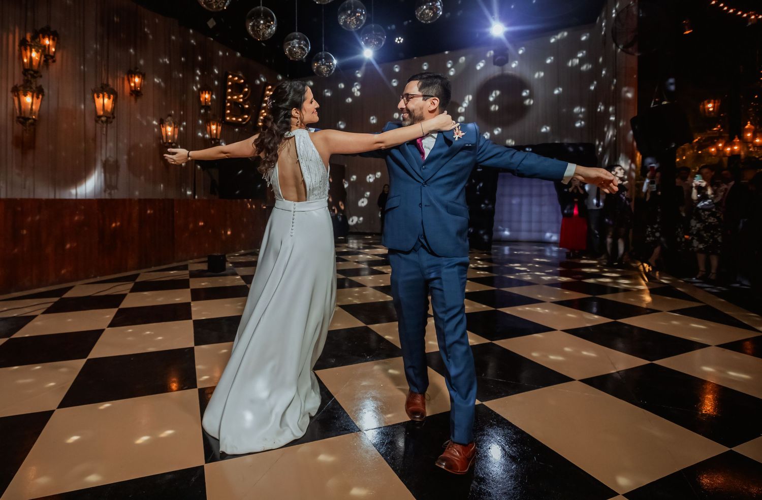 Bride and groom dance gracefully on checkered floor with glowing lanterns on wood wall.