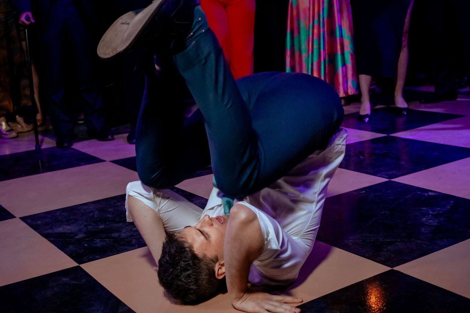 Wedding guest performs impressive breakdance move on checkered dance floor at energetic wedding reception party.