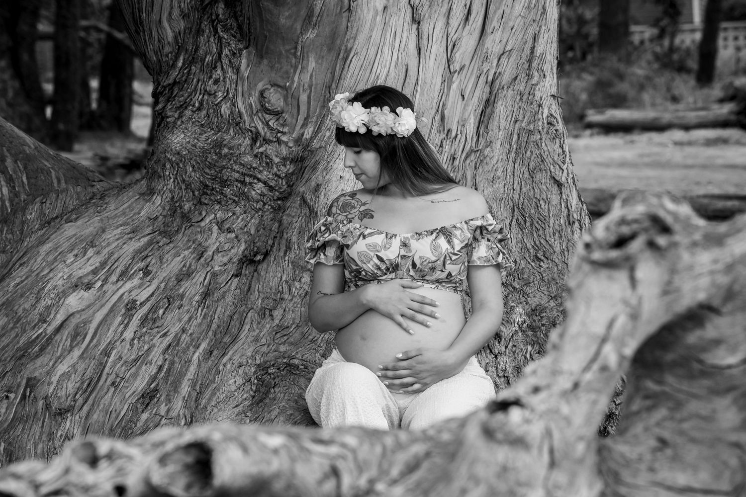 Black and white photo of pregnant woman with flower crown leaning against large tree.