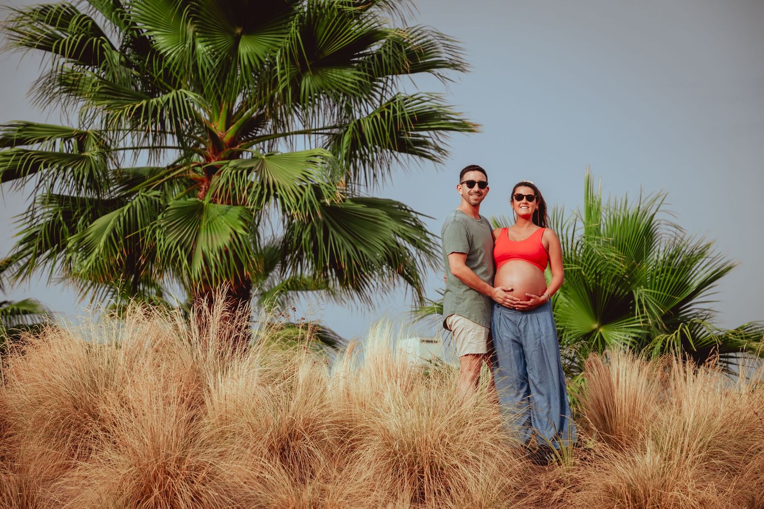 Couple standing together near palm trees, pregnant woman in red top with bare belly.