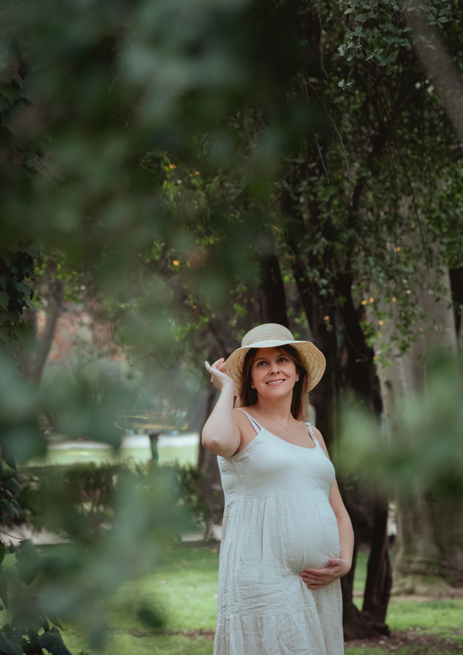 Smiling pregnant woman in white dress and straw hat standing among green trees in a park.