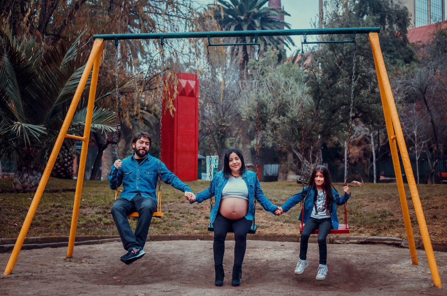 Family maternity photo with parents and child holding hands on colorful playground swings in park.